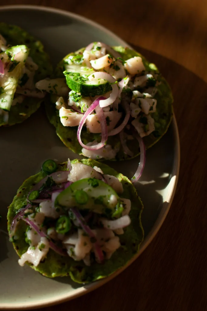 Países con mejor gastronomía: Tres tostadas verdes cubiertas con ceviche fresco, hechas de pescado blanco, rodajas de pepino, cebolla morada, chiles verdes y cilantro, presentadas en un plato bajo la luz del sol